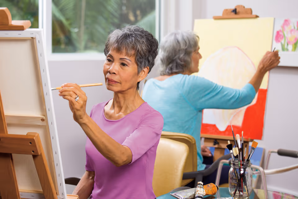Two elderly women painting on canvases set on easels in a bright room with a window showing greenery outside. One woman in a purple shirt is focused on her painting, while the other woman in a blue shirt is painting a floral picture. A jar with paintbrushes and tubes of paint are on the table in front of them.