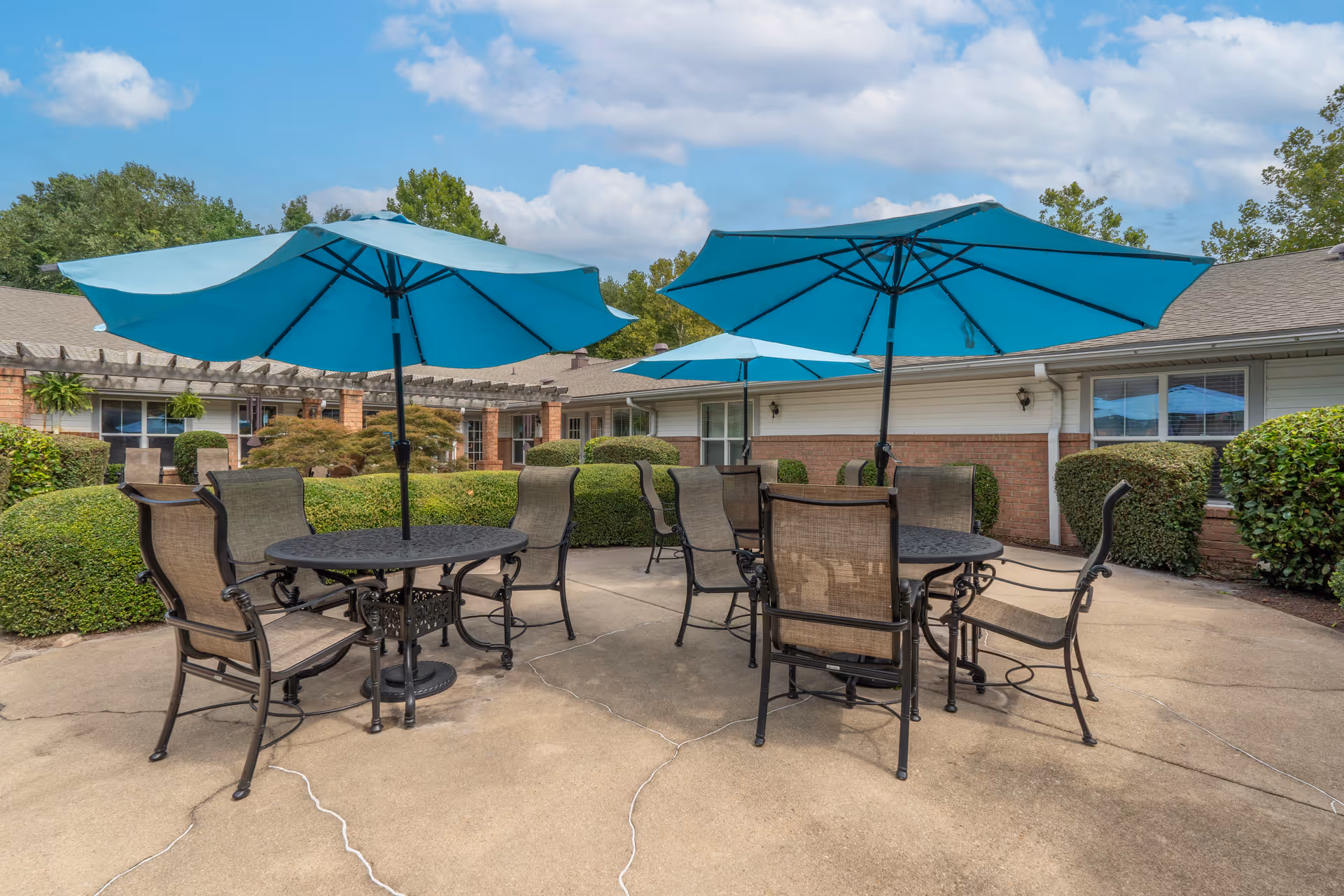 Outdoor patio area with multiple round metal tables and chairs, each table shaded by large blue umbrellas. The patio is surrounded by trimmed bushes and a single-story brick building under a partly cloudy sky.