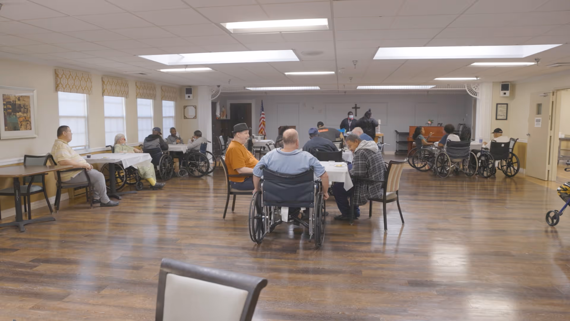 A large room with wooden flooring and several elderly individuals, many in wheelchairs, seated around tables and along the walls. The room has multiple windows with patterned valances, fluorescent ceiling lights, and an American flag in the background. The setting appears to be a communal area in a healthcare or rehabilitation facility.