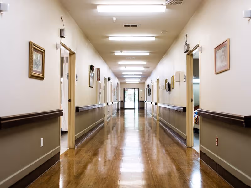 Long, polished hallway inside a nursing facility with handrails, open resident room doors, framed artwork, and fluorescent ceiling lights.