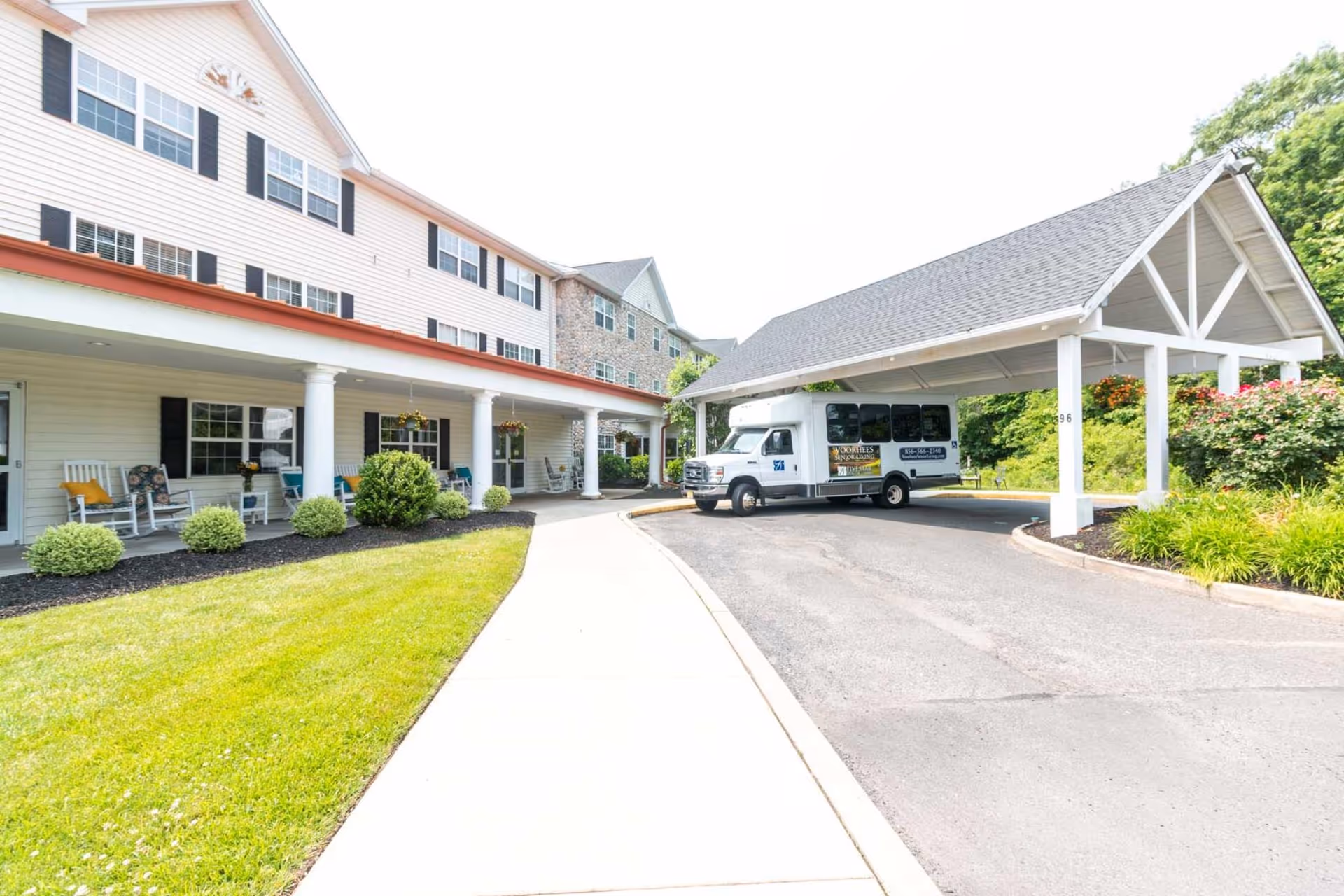 Exterior view of a senior living facility with a covered entrance area where a white shuttle bus is parked. The building has white siding, black shutters, and a porch with rocking chairs and potted plants. There is a well-maintained lawn and landscaped bushes along the sidewalk leading to the entrance.