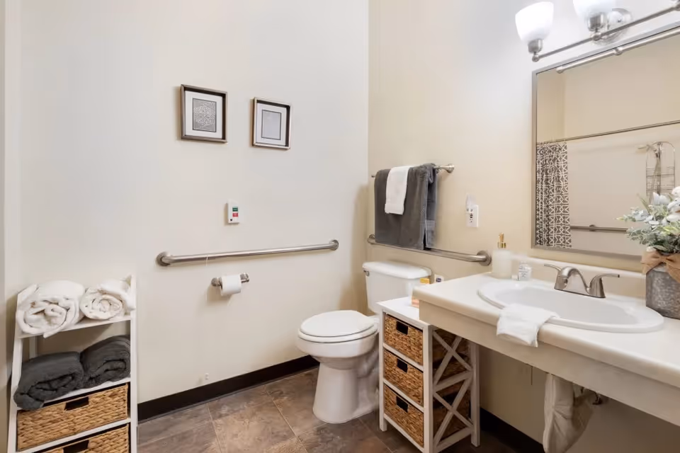 A clean and accessible bathroom with a white toilet, a white sink with a large mirror above it, and a small shelving unit holding rolled towels and baskets. There are grab bars on the walls near the toilet and sink, two framed pictures on the wall, and a towel rack with gray and white towels. The floor is tiled and the walls are painted a light beige color.