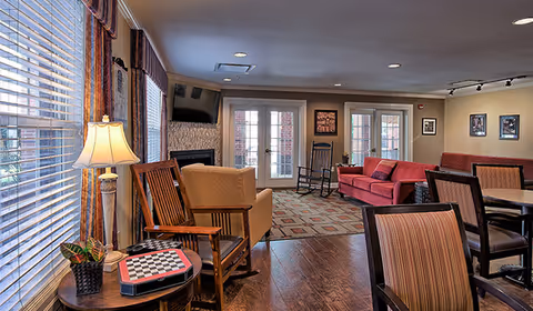 Bright communal living room with chairs, sofas, a fireplace and TV facing French doors and large windows.