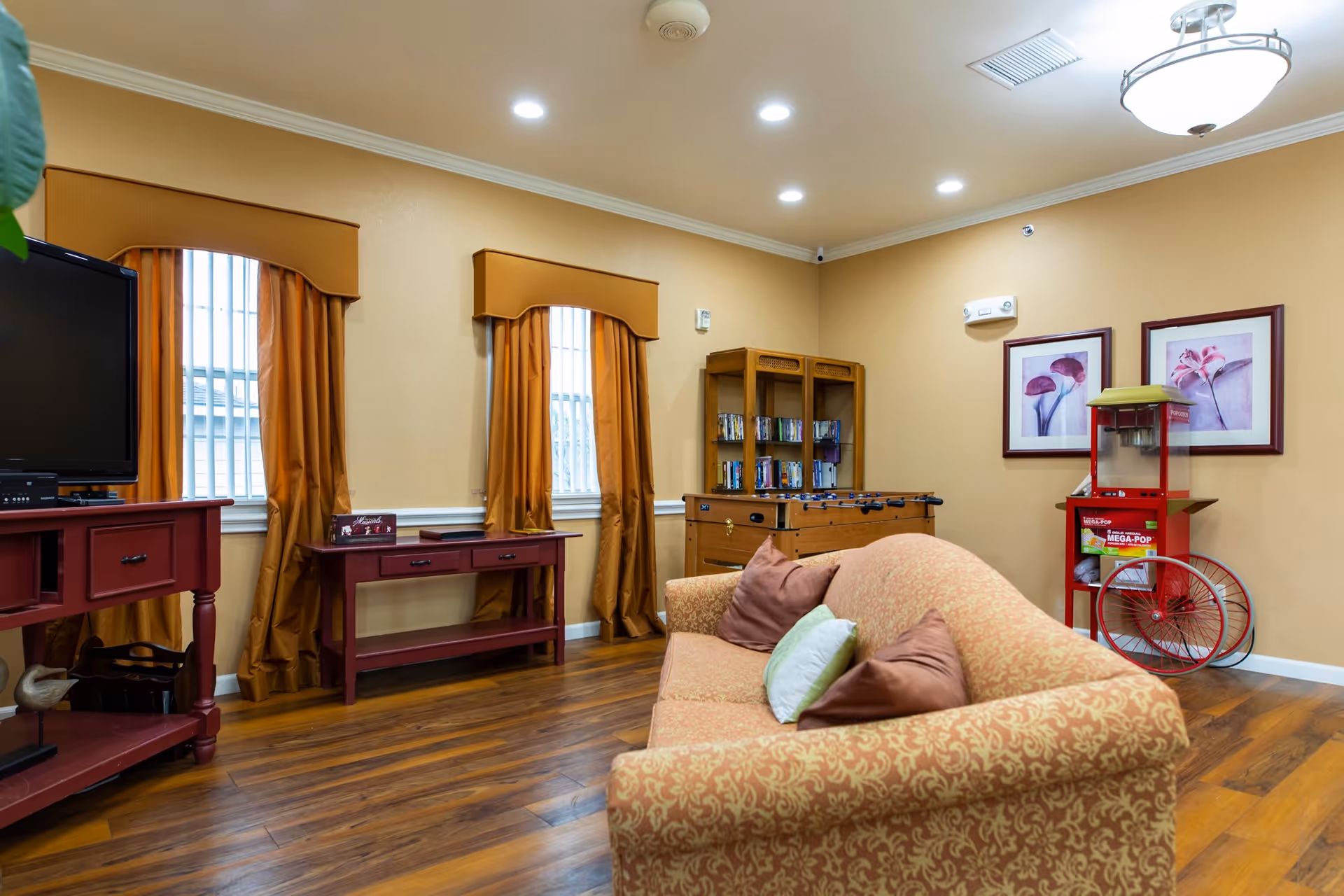 A cozy living room area with a patterned orange sofa adorned with brown and green pillows. The room features wooden flooring, two windows with orange curtains, a red wooden TV stand with a flat-screen TV, a matching red console table, a foosball table, a bookshelf with books, and a red popcorn machine. The walls are painted beige and decorated with two framed floral pictures.