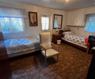 A small bedroom with two single beds placed against opposite walls, each with bedding and pillows. A white cushioned chair is positioned in the center of the room on a brown carpet. There is a wooden nightstand with a lamp and some items between the beds. The walls are white with two windows covered by blinds and curtains, and a framed picture hangs on the wall. A mirror is mounted above the nightstand.