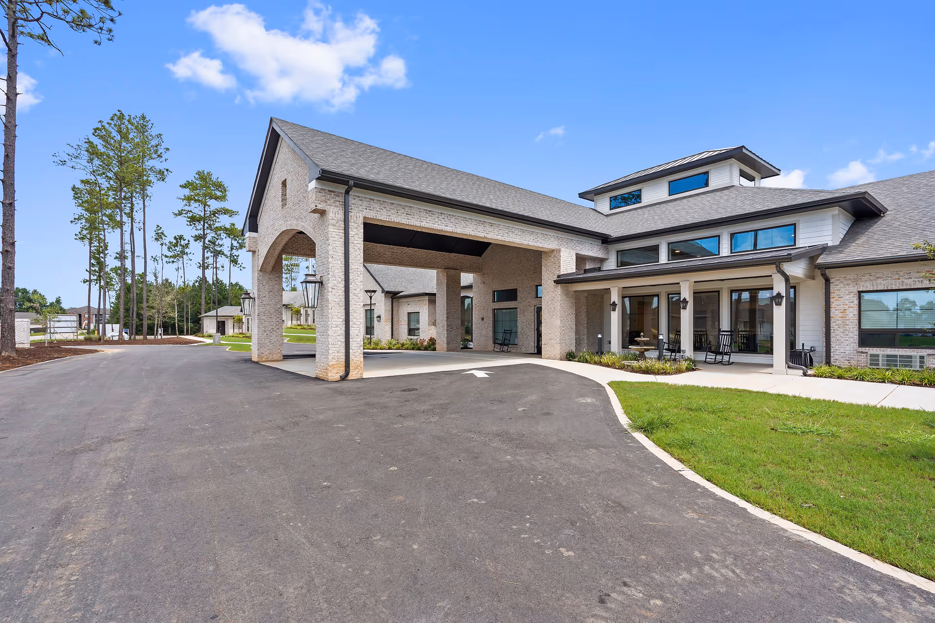 Exterior view of a senior living facility named Sage Lake featuring a covered entrance with brick pillars, a paved driveway, green lawn, and tall pine trees under a blue sky with some clouds.