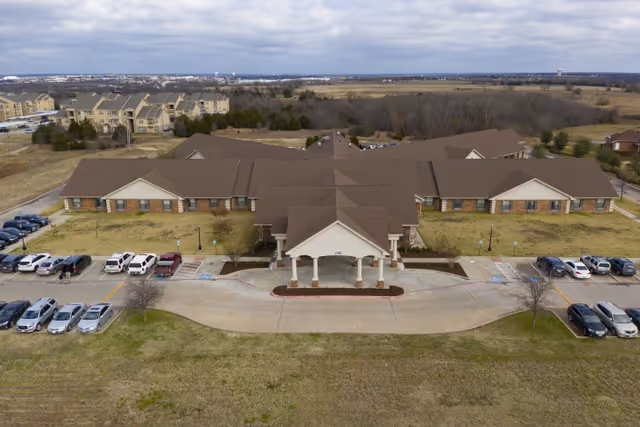 Aerial view of a single-story healthcare and rehabilitation facility with a large covered entrance and multiple wings extending from the center. Several cars are parked in front of the building, and the surrounding area includes open grassy fields and some trees under a cloudy sky.