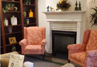 A cozy interior living area featuring two patterned pink armchairs positioned around a white fireplace with decorative items on the mantel. To the left, there is a dark wooden bookshelf with various decorative objects and plants. A small table with papers and a certificate is partially visible in the foreground.