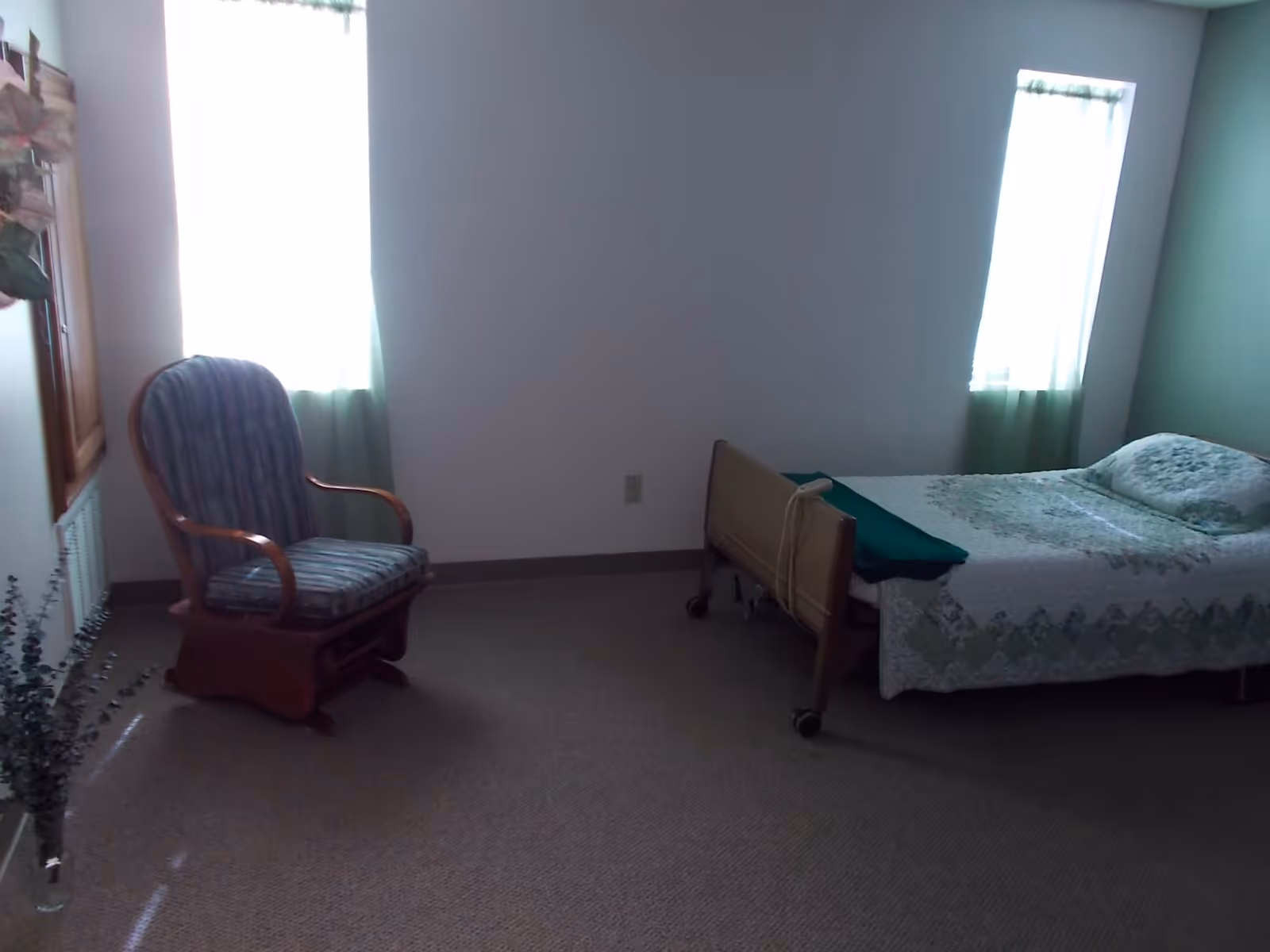 Simple assisted-living bedroom with a single adjustable bed, patterned bedspread, and a cushioned wooden armchair beside two windows.