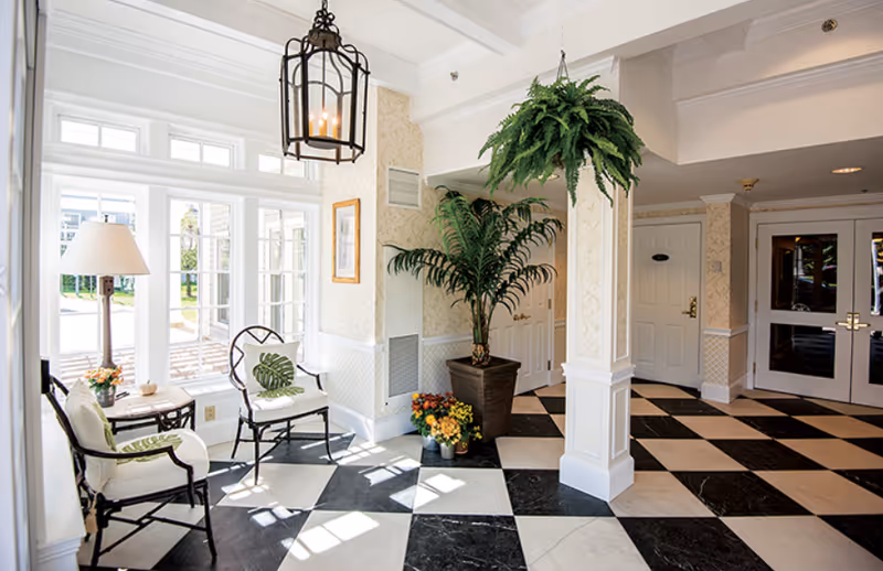 Bright and airy interior space with black and white checkered floor tiles, white walls with decorative molding, and large windows letting in natural light. The room features two cushioned chairs with green leaf-patterned pillows, a small side table with a lamp and flowers, a large potted plant, a hanging fern, and double doors in the background.