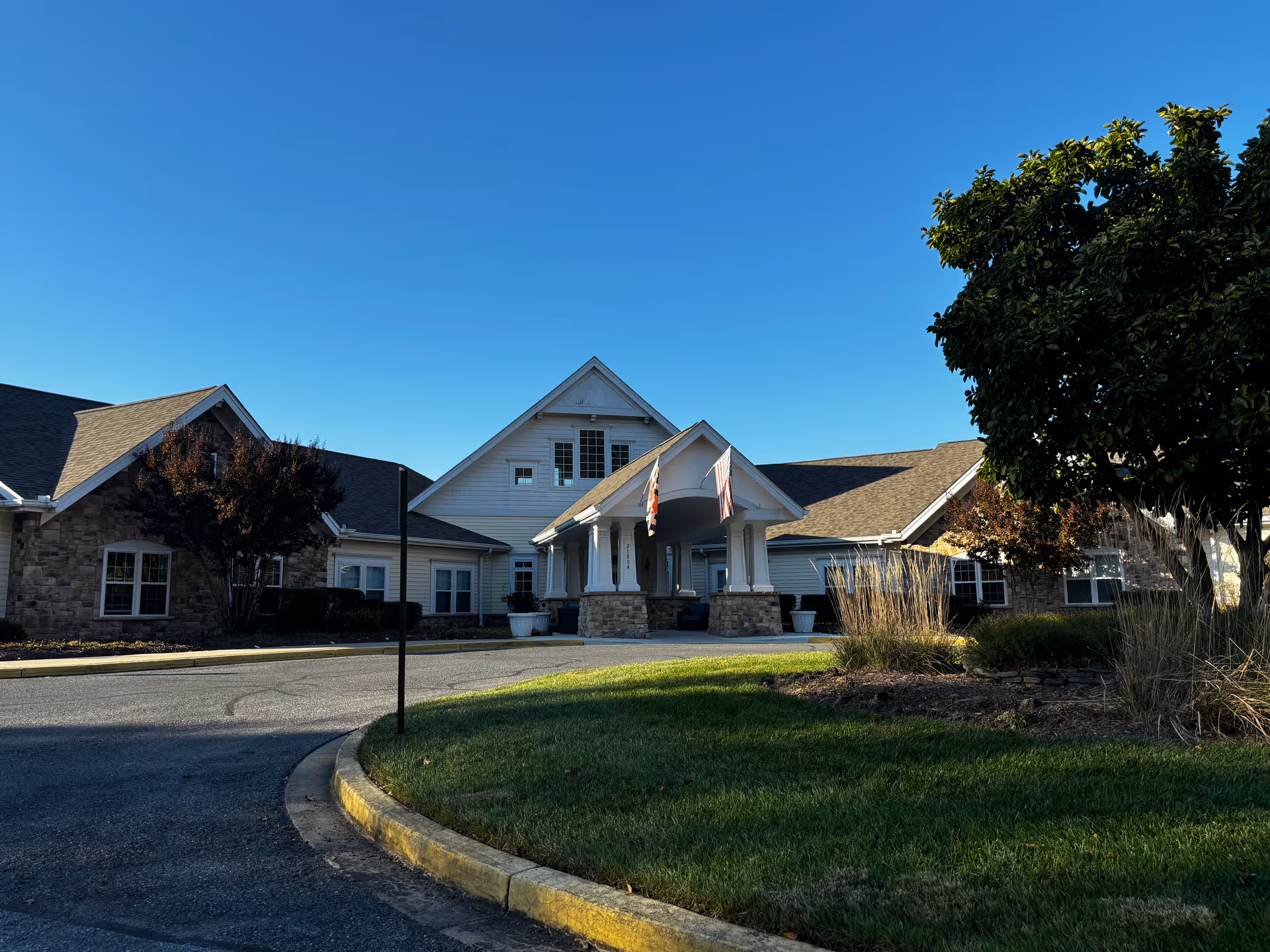 Exterior view of Somerford House Frederick, showing a single-story building with a covered entrance supported by white columns. The building has a mix of stone and siding on the facade, with several windows and a peaked roof. There are two flags hanging from the entrance roof, and the surrounding area includes a curved driveway, green grass, and trees under a clear blue sky.
