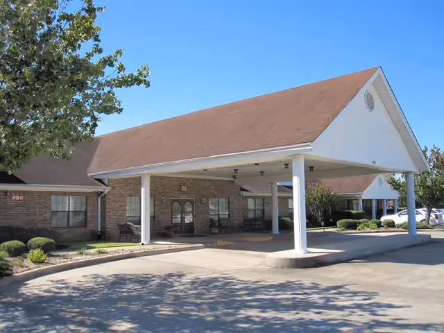 Exterior view of Cambridge Square Assisted Living facility showing a covered entrance with white pillars, brick walls, and a brown sloped roof under a clear blue sky. There are some bushes and trees around the building and a parking area with cars visible in the background.