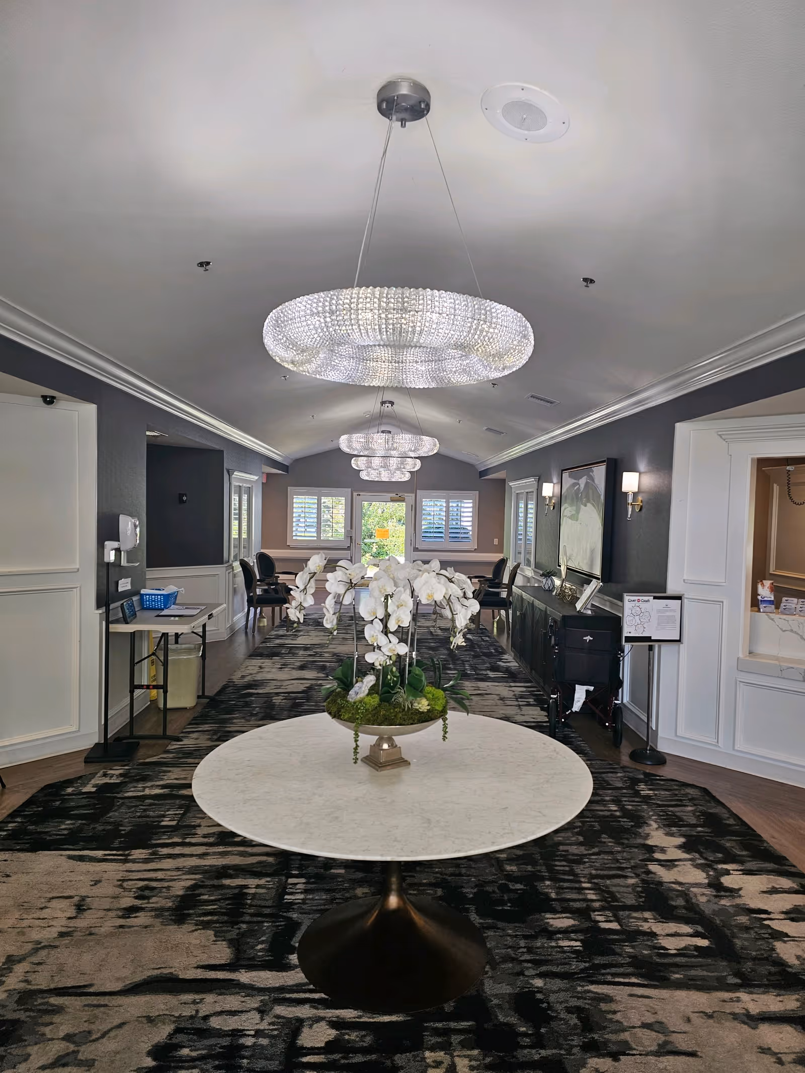 Interior hallway of Quartz Hill Post Acute facility featuring a round table with a floral arrangement in the center, modern chandeliers hanging from the ceiling, and seating areas with chairs and tables along the sides near windows with white shutters.