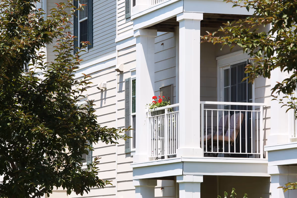 Exterior of a light-colored residential building showing a small balcony with chairs and a flower box, partially obscured by trees.