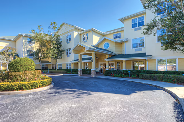 Exterior view of a three-story senior living facility building with a covered entrance, surrounded by landscaped bushes and trees under a clear blue sky.