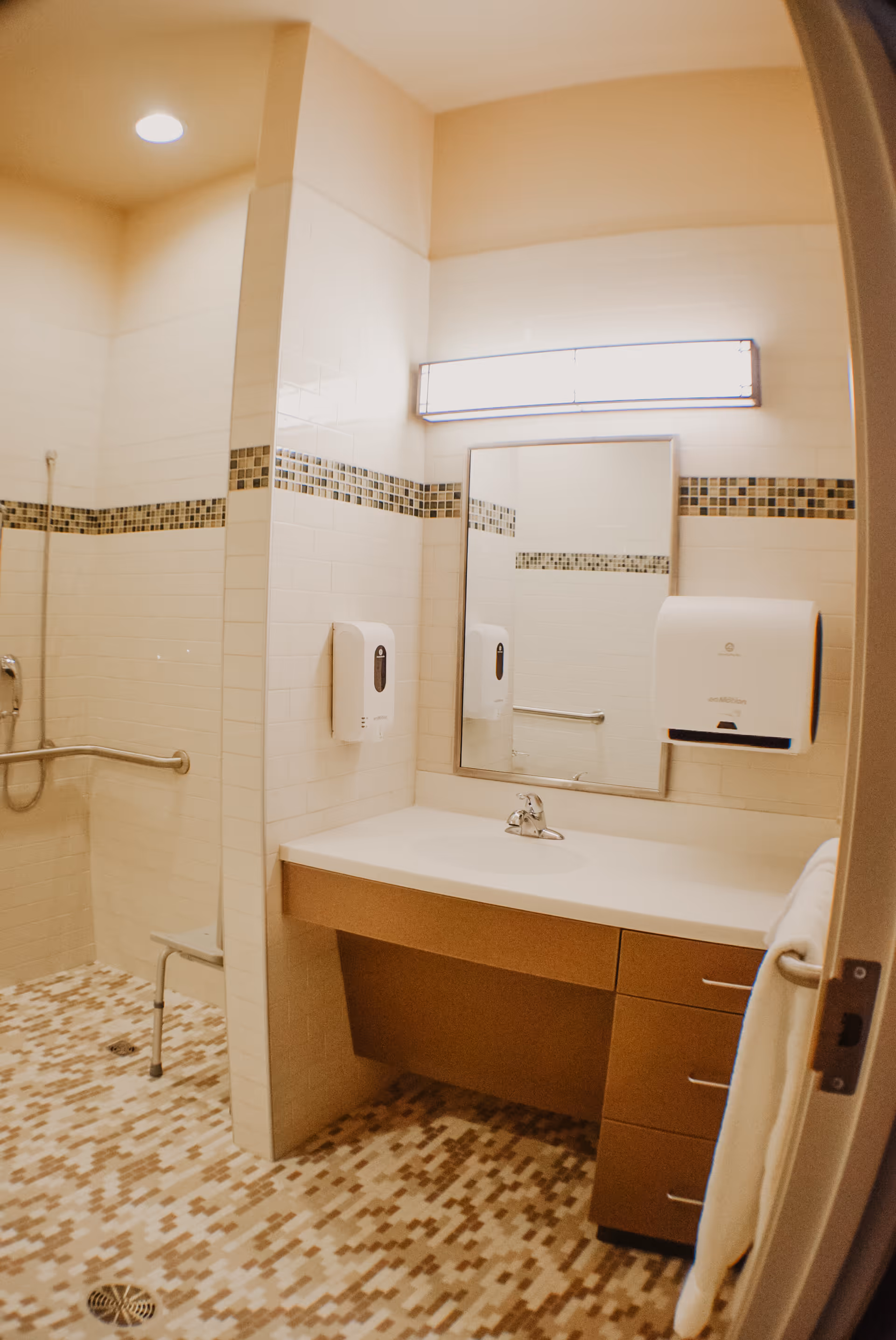 A clean and accessible bathroom with a walk-in shower featuring a handheld showerhead and grab bars. The bathroom has a tiled floor with a mosaic pattern, white tiled walls with a decorative horizontal strip of small square tiles, a large mirror above a sink with a countertop, a soap dispenser, and a paper towel dispenser. A towel hangs on a bar attached to the door.