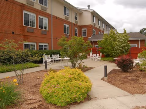 Outdoor courtyard area of a senior living facility with paved walkways, green shrubs, small trees, and white benches. The building has red brick and beige siding with multiple windows.