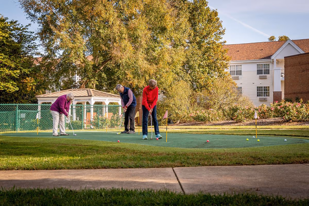 Three older adults playing on a putting green outside a residential building surrounded by trees and lawn.