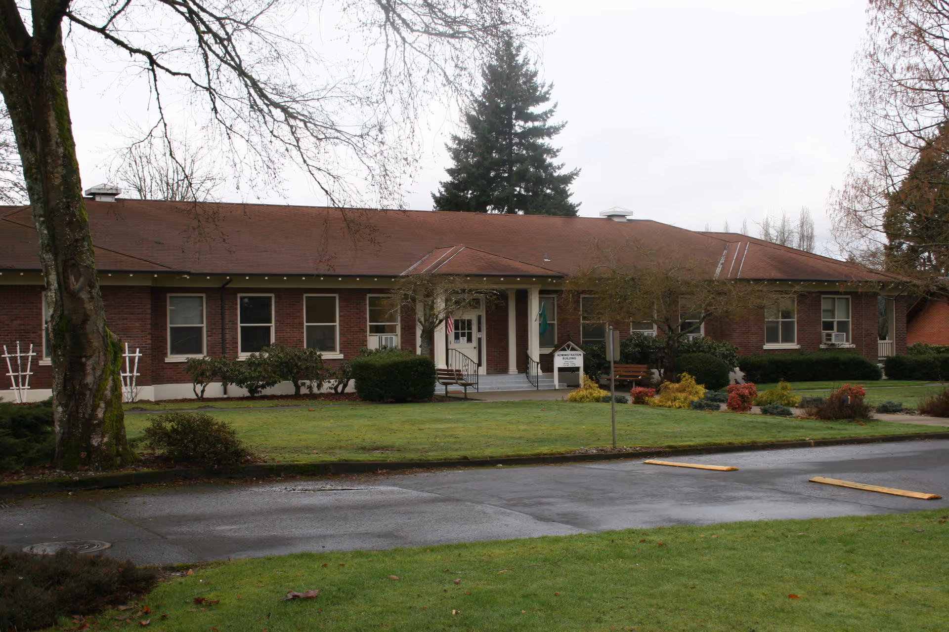 Exterior view of a single-story brick building with a brown roof, surrounded by a well-maintained lawn and trees. There is a small entrance with steps and a handrail, a bench near the entrance, and a signpost in front of the building. The sky is overcast.