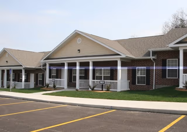 Exterior view of a single-story brick building with beige siding and a gabled roof, featuring white columns and railings on the front porch. There is a paved parking lot with marked parking spaces in front of the building.