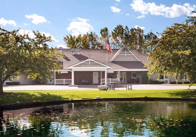 Single-story retirement facility building with a covered entrance, an American flag, and a reflective pond in the foreground.