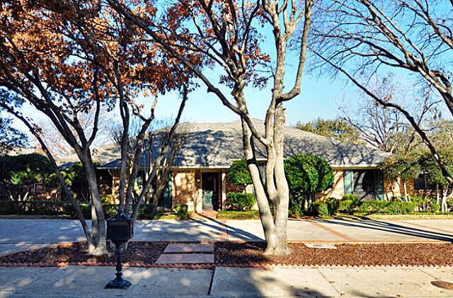 Single-story residential building with a sloped roof, surrounded by leafless trees and shrubs, with a paved driveway and sidewalk in front under a clear blue sky.