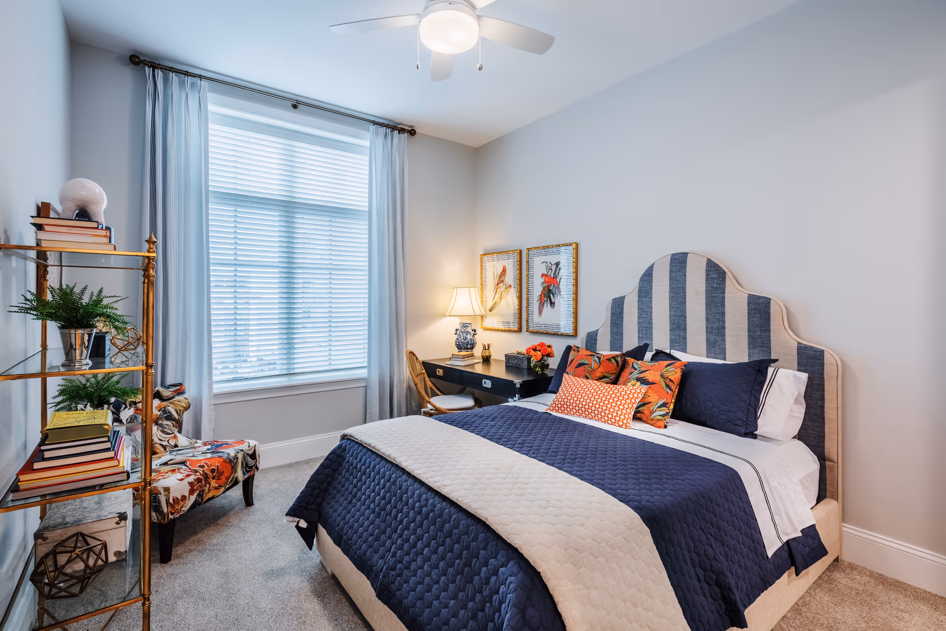 Bright, neatly staged bedroom with a striped upholstered headboard, navy-and-white bedding, a desk, decorative shelving and a window with sheer curtains.