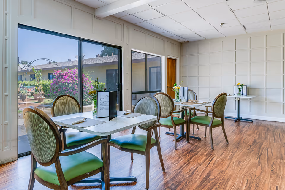 A bright dining room with wooden floors, several tables with green cushioned chairs, menus, and small flower vases on each table. Large sliding glass doors and windows provide a view of a garden with blooming flowers outside.