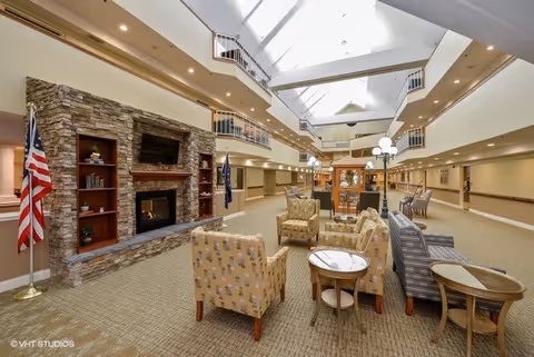 Spacious and well-lit common area in a senior living facility featuring a stone fireplace with built-in shelves, multiple seating arrangements with armchairs and small tables, American and state flags, and a high ceiling with skylights and balconies overlooking the area.