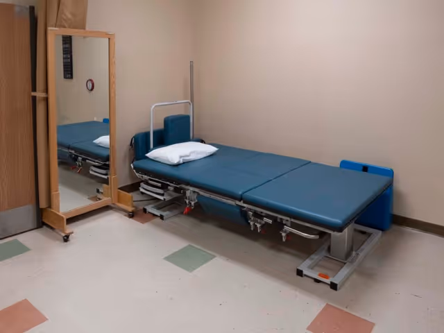 A medical examination room with a blue padded examination table featuring a white pillow. A full-length mirror on a wooden frame with wheels is positioned next to the table. The room has beige walls and a tiled floor with green and brown square patterns.