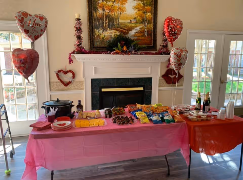 A festive snack table set up in a room with a fireplace and large windows. The table is covered with pink and red tablecloths and holds various snacks including chips, cheese, strawberries, cupcakes, and bottles of sparkling cider. Heart-shaped balloons and Valentine's Day decorations adorn the area, with a painting hanging above the fireplace.