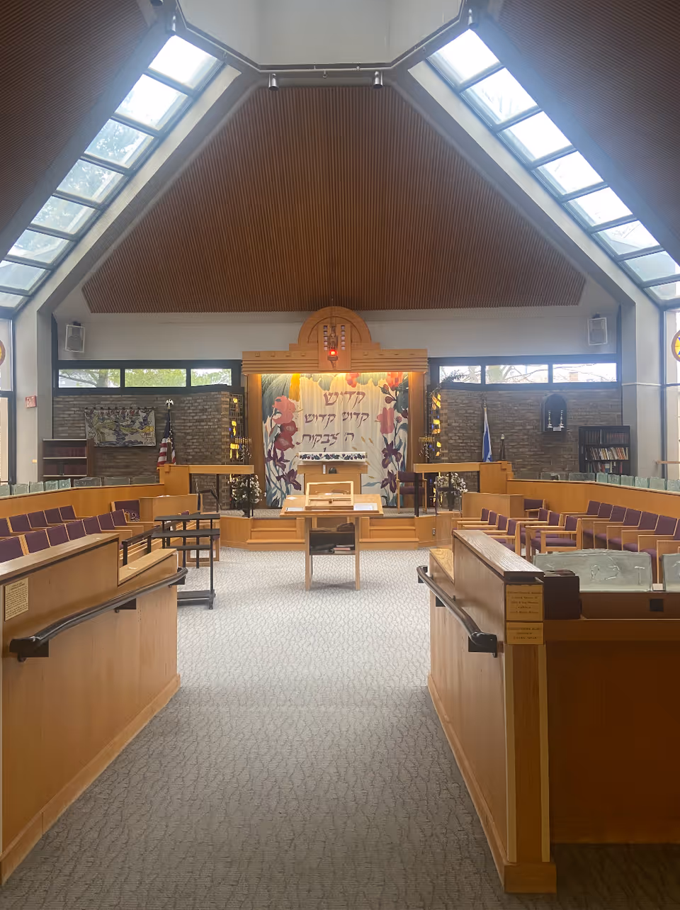 Interior of a synagogue-style chapel with a wood-paneled ceiling, rows of seating, a central aisle, and an ark with a Hebrew tapestry at the front.