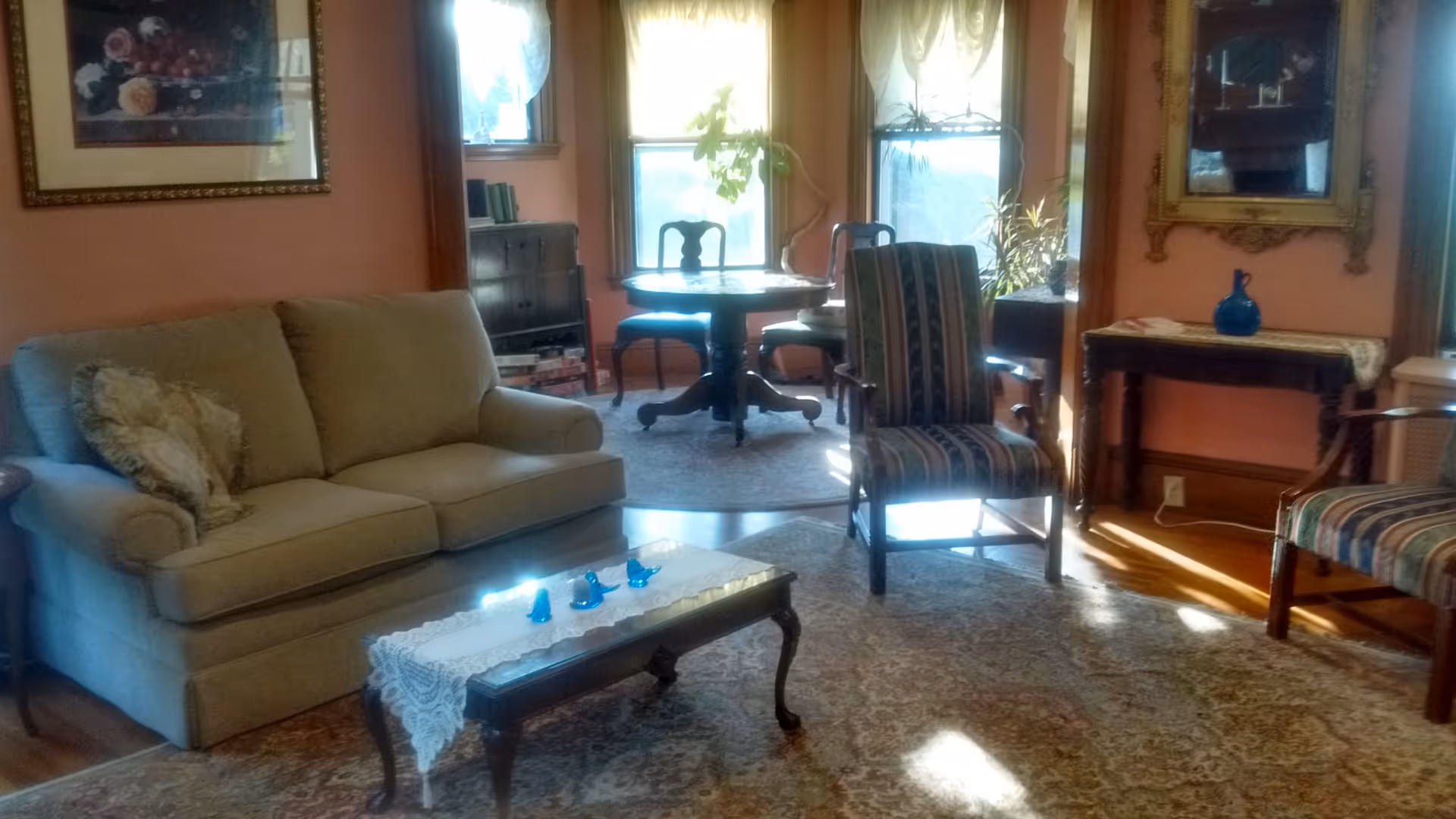 A cozy living room with a beige sofa, two striped armchairs, a wooden coffee table with a lace runner and small blue decorative items, a round dining table with chairs near three windows, a framed floral painting on the wall, and a large ornate mirror above a small wooden table with a blue vase.