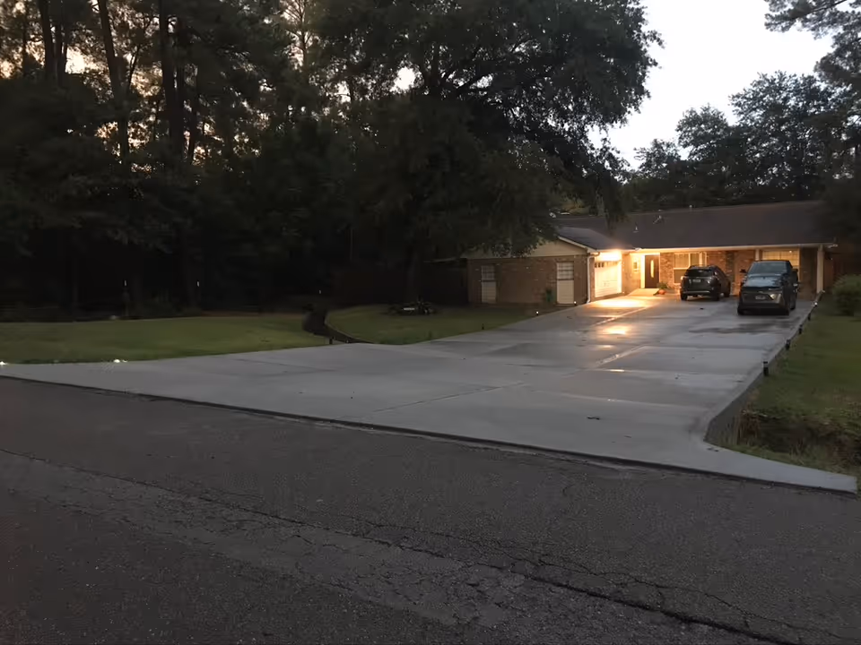 A single-story brick building with a long driveway and two parked cars near the entrance, surrounded by trees and grass, taken during dusk with lights on near the entrance.