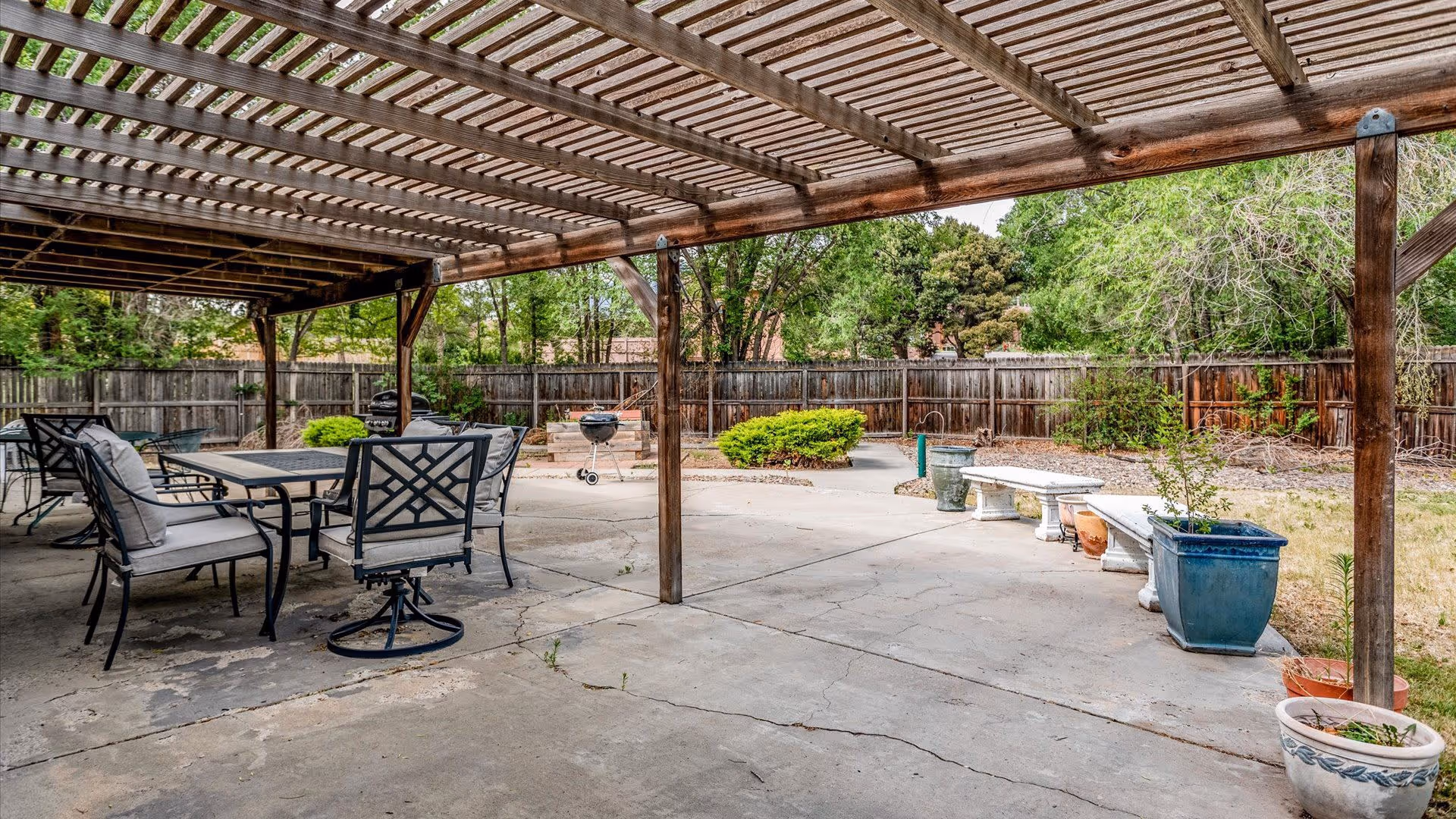 Shaded concrete patio with a wooden pergola, outdoor dining set, planters, and a fenced backyard.