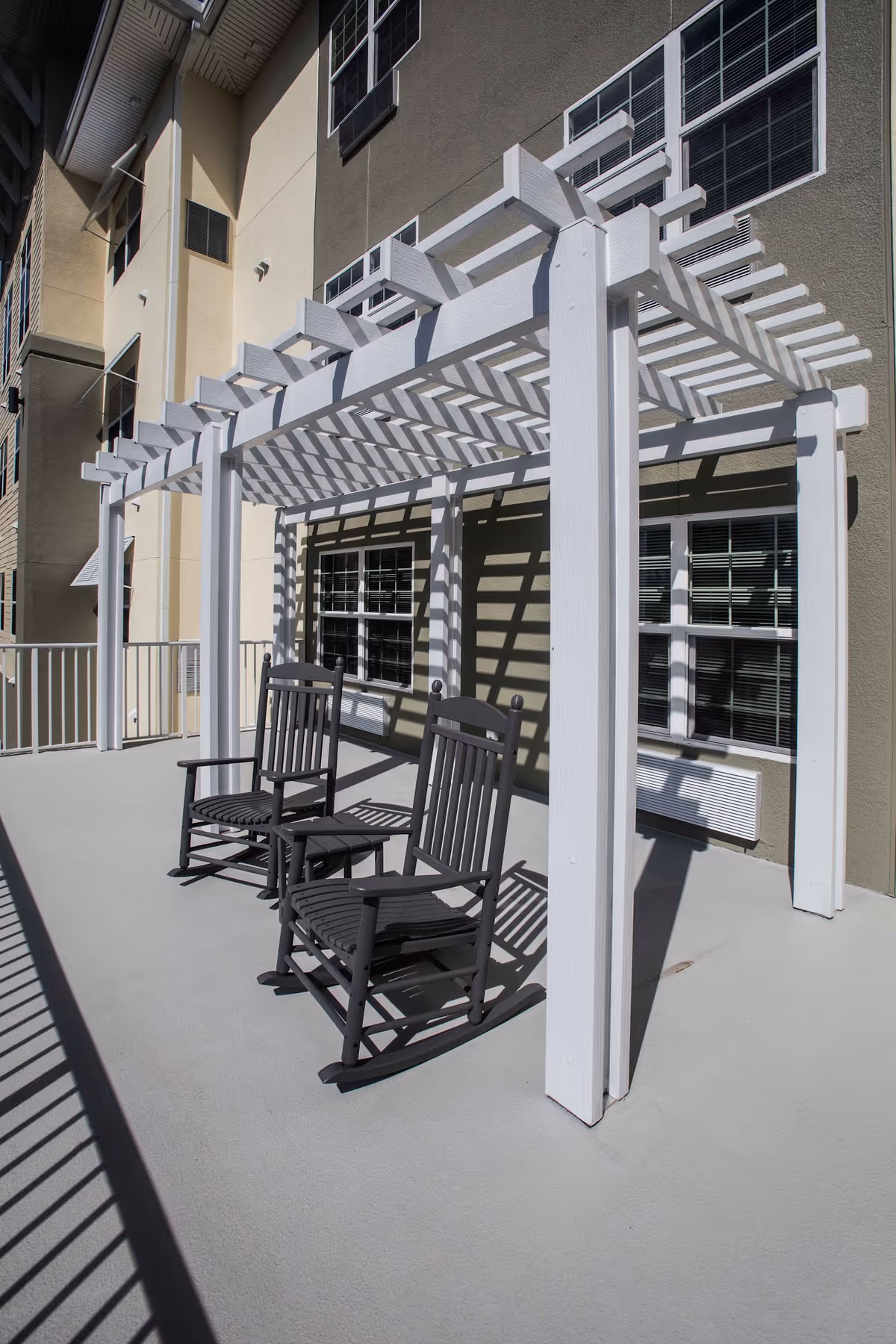 Outdoor patio area with two black rocking chairs under a white pergola attached to the side of a beige building with multiple windows.