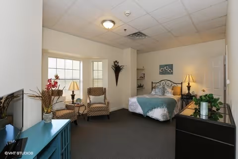 A well-lit bedroom in an assisted living facility featuring a bed with white and teal bedding, two patterned armchairs near a window with a lamp on a small table between them, a teal cabinet with decorative items, a black dresser with plants, and neutral-colored walls and carpet.