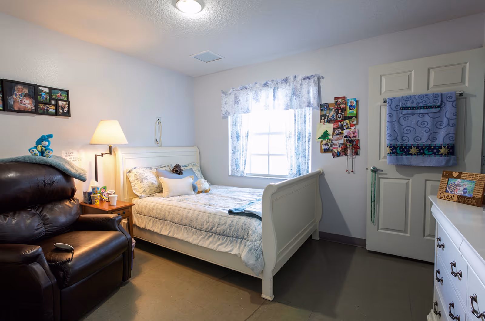 A cozy bedroom with a white wooden bed covered in light blue and white bedding, a brown leather recliner chair with a blue stuffed bunny on it, a wooden nightstand with a lamp and cups, a window with floral curtains, a door with towels hanging on it, and a white dresser with a photo frame and decorations.