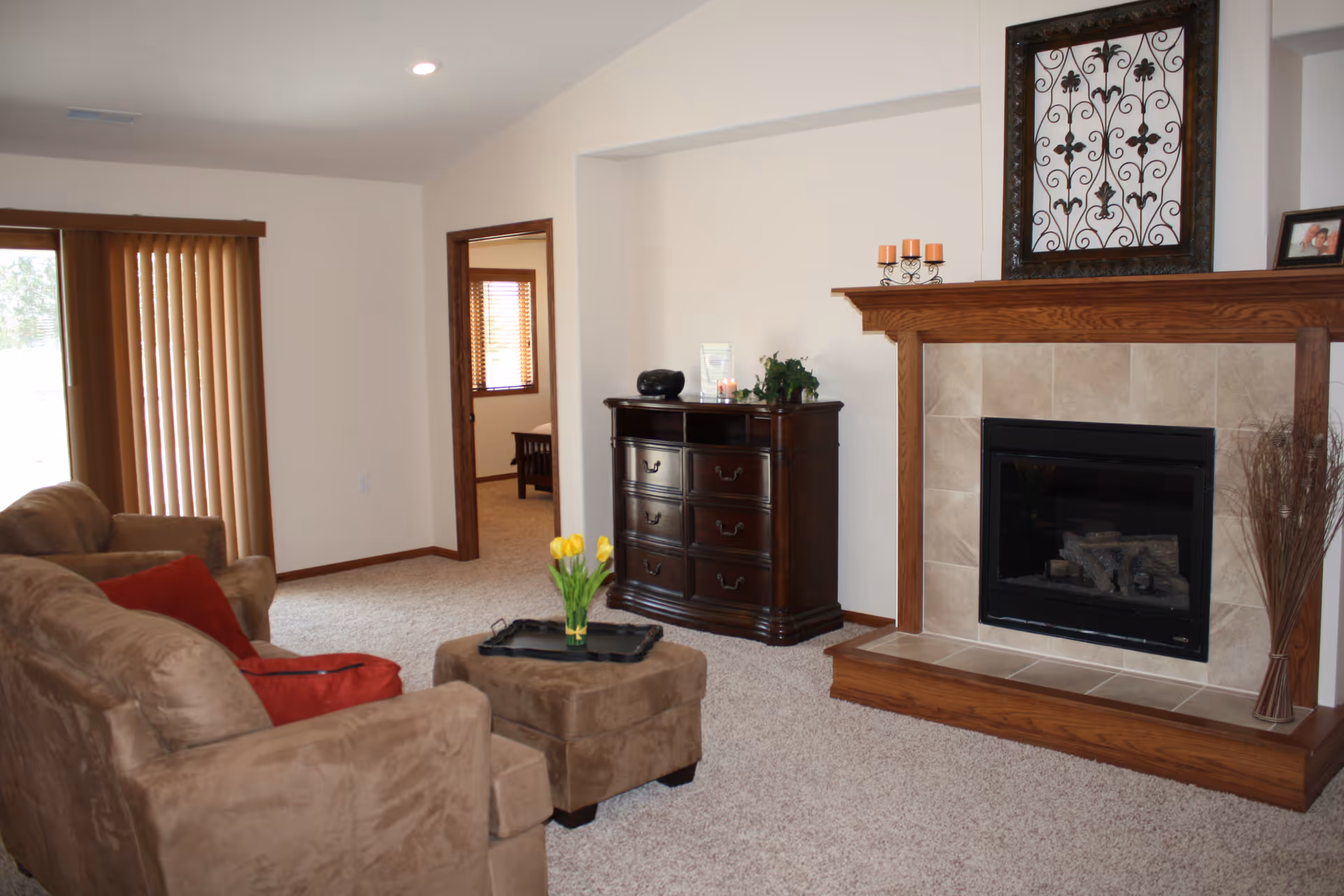 A cozy living room with beige carpet, a brown sofa and armchair with red cushions, a matching ottoman with a tray and yellow tulips, a wooden dresser, and a fireplace with a wooden mantel decorated with candles and framed artwork.