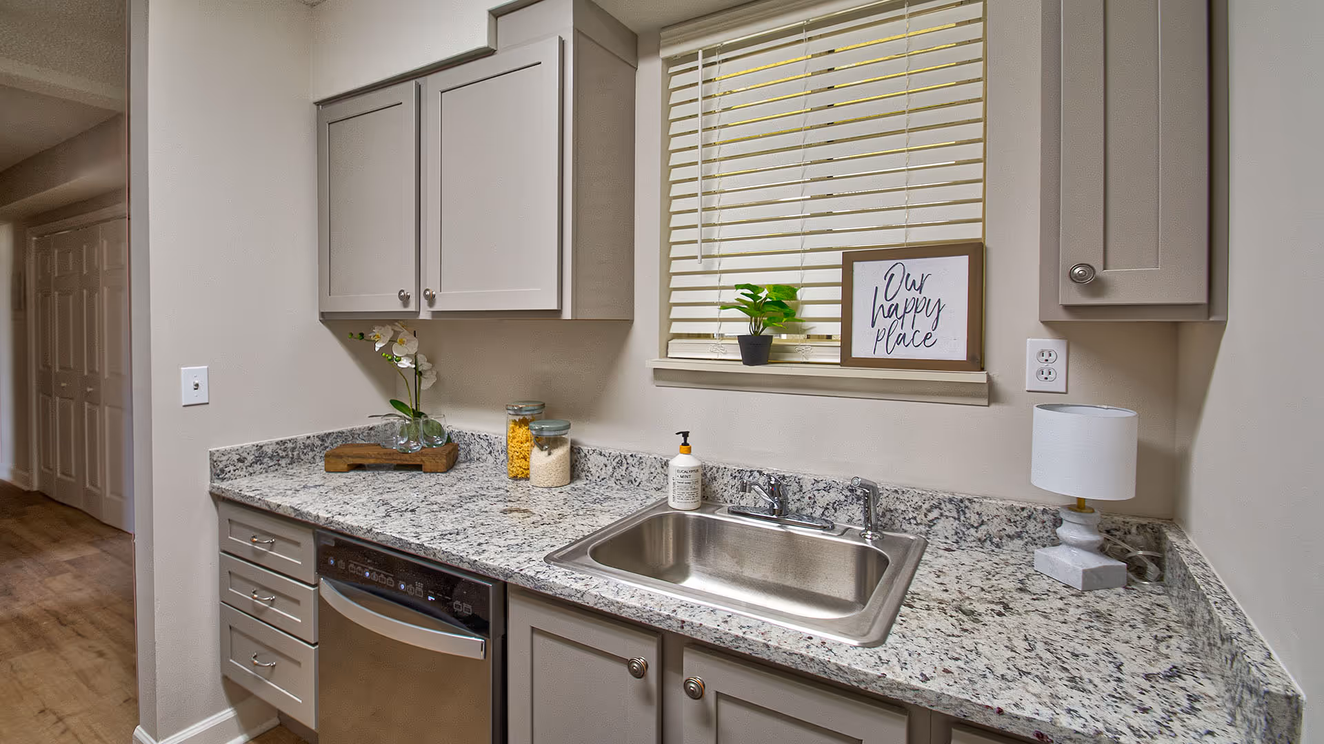 A clean kitchen countertop with a stainless steel sink, a small lamp, a soap dispenser, and decorative items including a small plant and a framed sign that reads 'Our happy place'. There are light gray cabinets above and below the countertop, and a window with closed blinds above the sink.