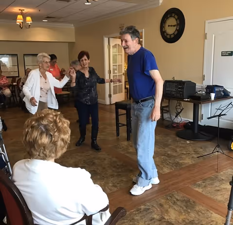 A group of elderly people dancing and enjoying themselves in a common room with wooden flooring. One man in a blue shirt and jeans is dancing while two women hold hands and smile. Another person is seated in the foreground watching the activity. The room has beige walls, a large clock, and a table with audio equipment.