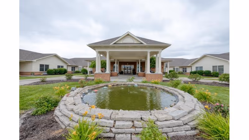 Covered front entrance and porte-cochere of a low-rise senior living building with a circular stone pond in the foreground.