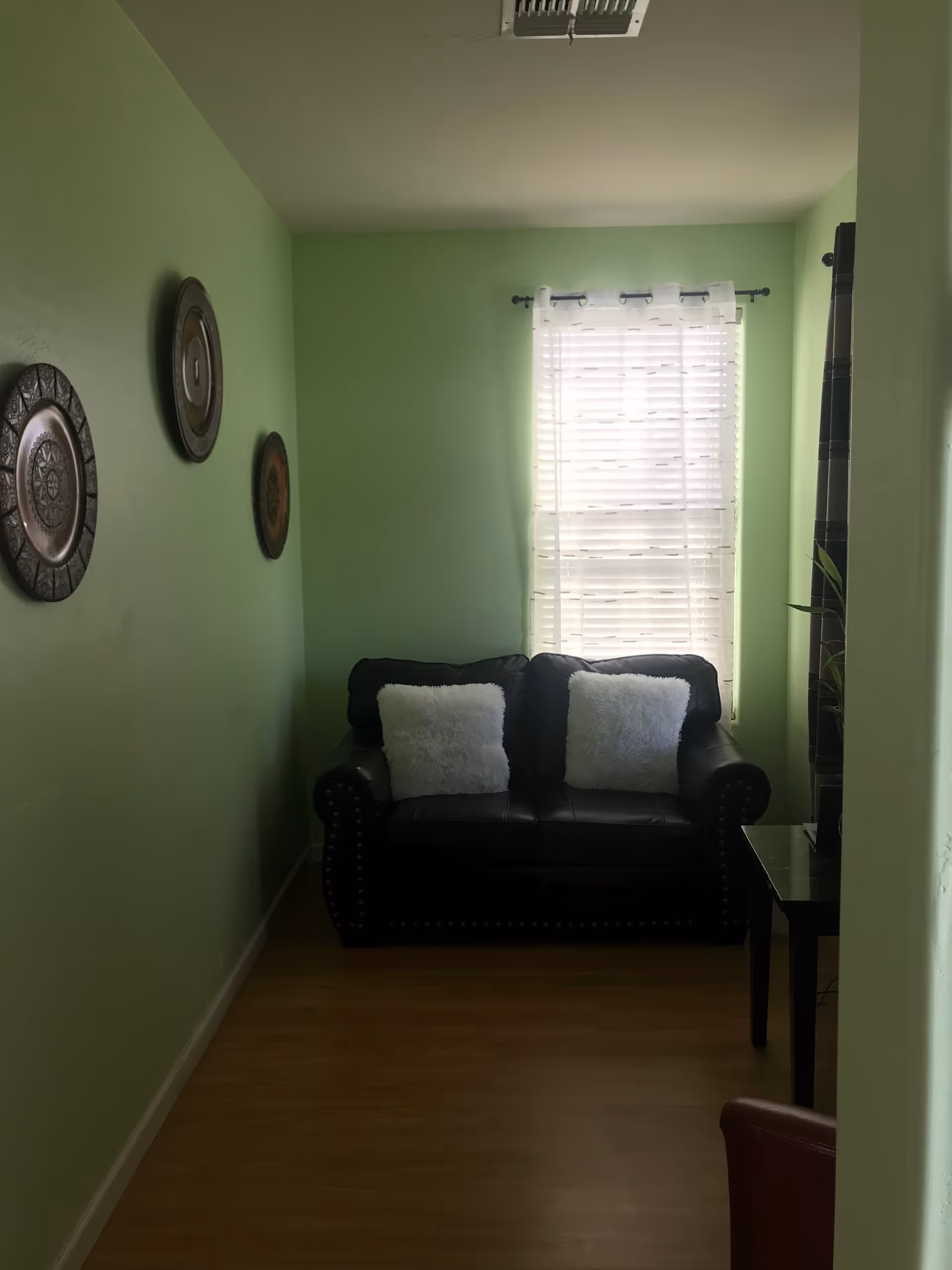 A small living room area with light green walls, a black leather loveseat with two white fluffy pillows, a window with white blinds and sheer curtains, three decorative round wall hangings on the left wall, and a small dark wooden side table on the right.