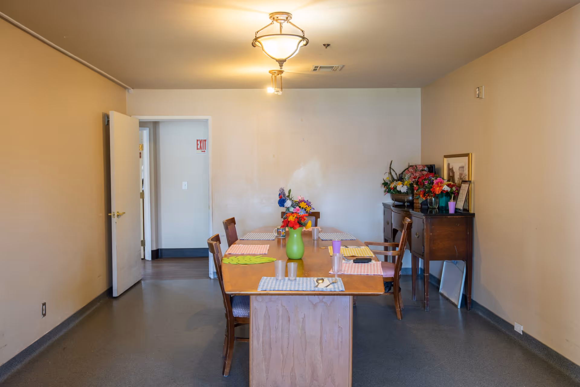 A dining room with a wooden table set with placemats, cups, and a green vase with colorful flowers. There are six wooden chairs around the table. Against the right wall is a wooden sideboard with more flower arrangements and framed pictures. The room has beige walls, a ceiling light fixture, and an open door leading to another area with an exit sign visible.
