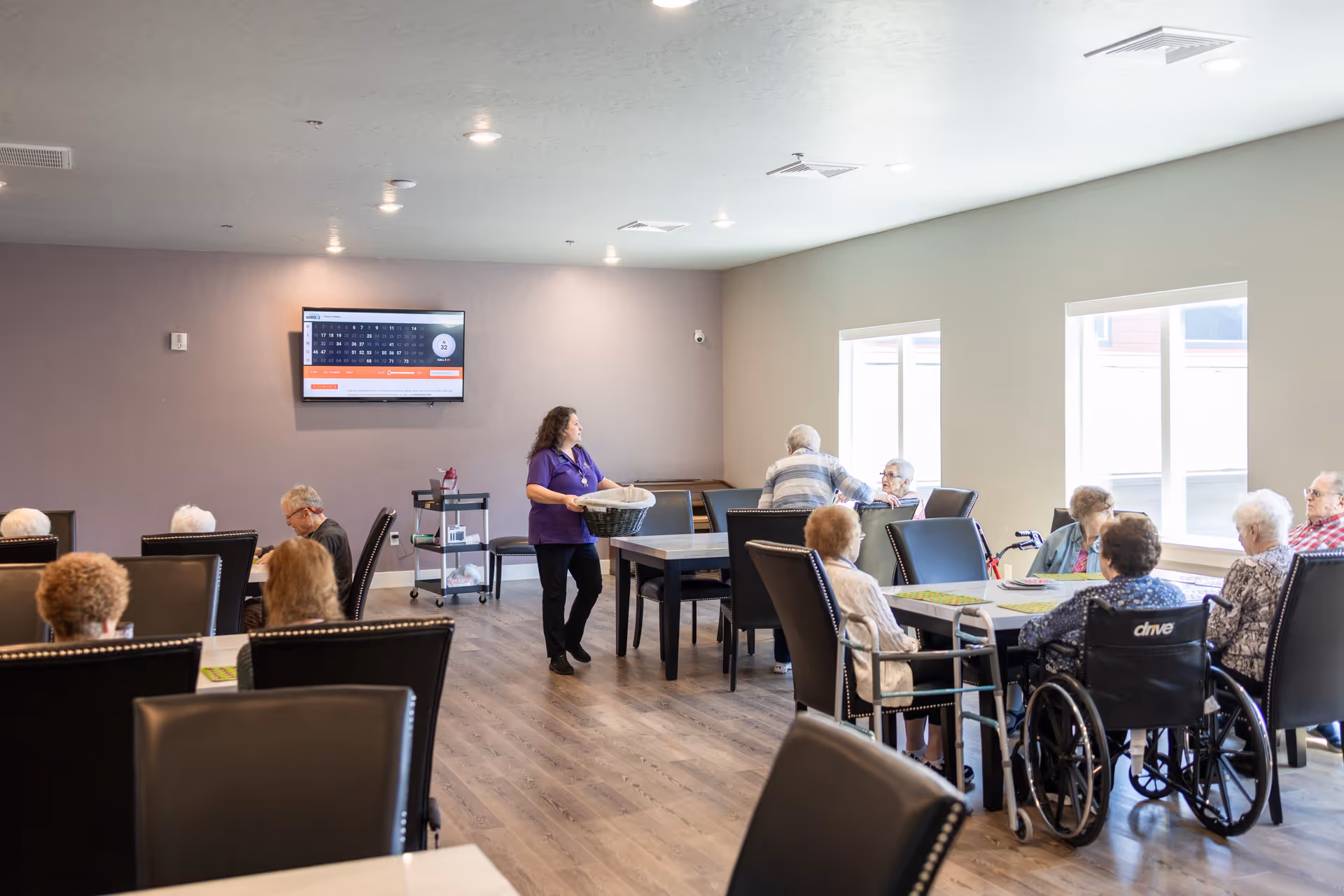 A group of elderly people sitting around tables in a well-lit room with large windows. A staff member in a purple shirt is standing and holding a basket. The room has wooden flooring, black cushioned chairs, and a TV mounted on the wall displaying a bingo game.