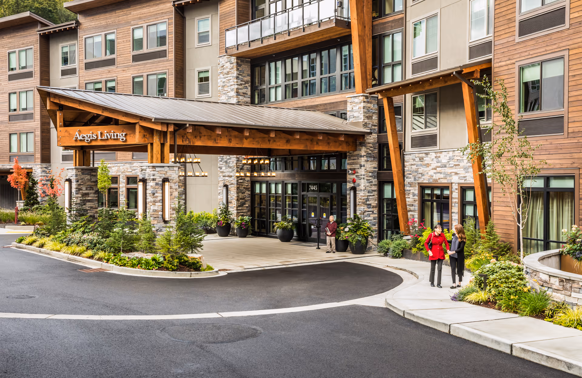 Exterior view of the Aegis Living Mercer Island senior living facility entrance with a covered drop-off area, stone and wood architectural details, landscaped greenery, and two women conversing on the sidewalk.