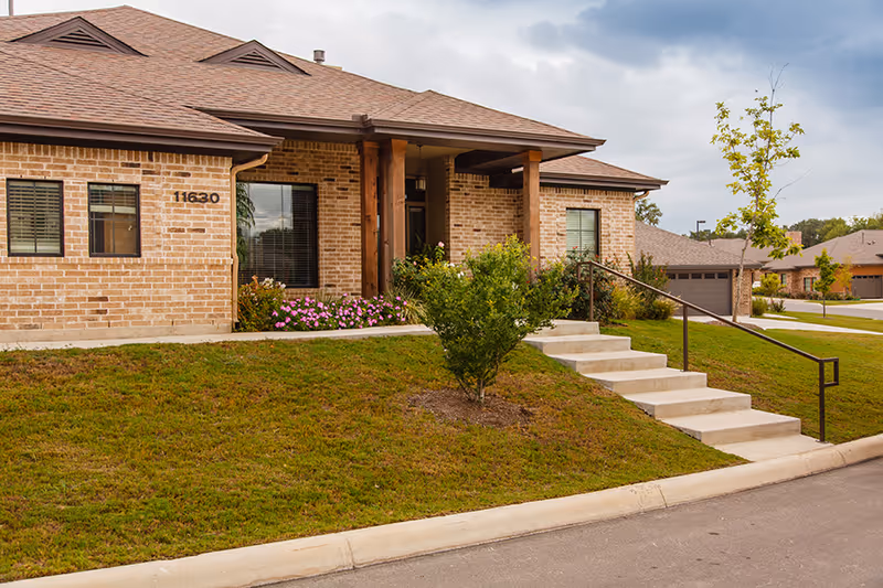 Exterior view of a single-story brick building with a sloped roof, a small porch supported by wooden columns, a set of concrete stairs with a metal handrail leading up to the entrance, and a well-maintained lawn with bushes and flowers.