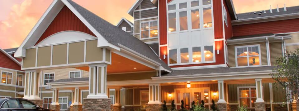Exterior view of a senior living facility building at sunset with a covered entrance supported by columns, multiple windows, and warm lighting inside.