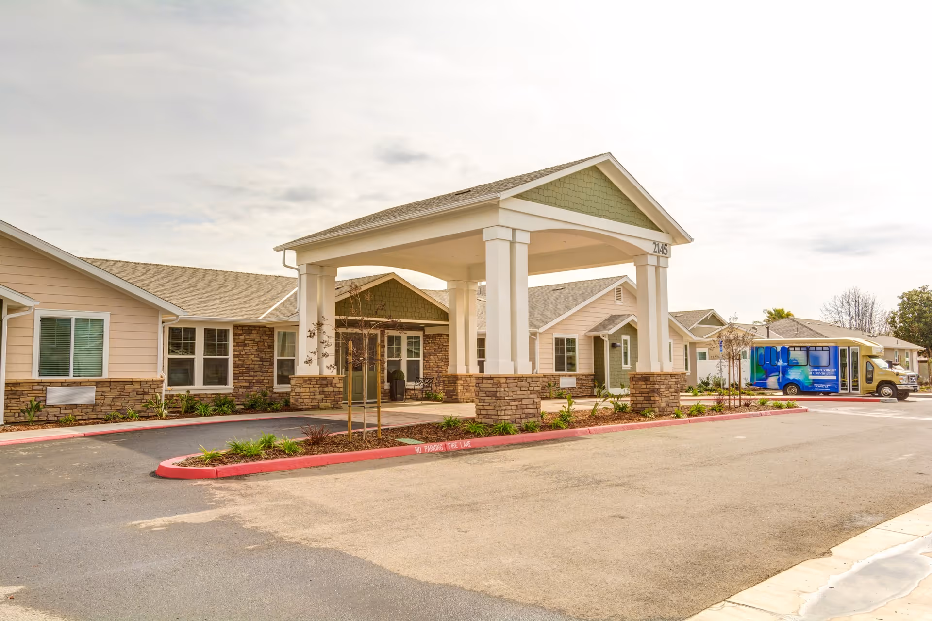 Exterior view of Carmel Village Memory Care facility showing a covered entrance with stone pillars and a driveway. The building has beige and green siding with multiple windows. A blue and white shuttle bus is parked on the right side near the building.