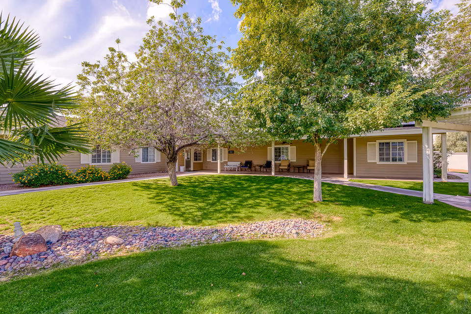 A well-maintained outdoor garden area at Paradise Valley Senior Living featuring green grass, two leafy trees, a rock bed, and a beige building with white window shutters and a covered porch with seating.